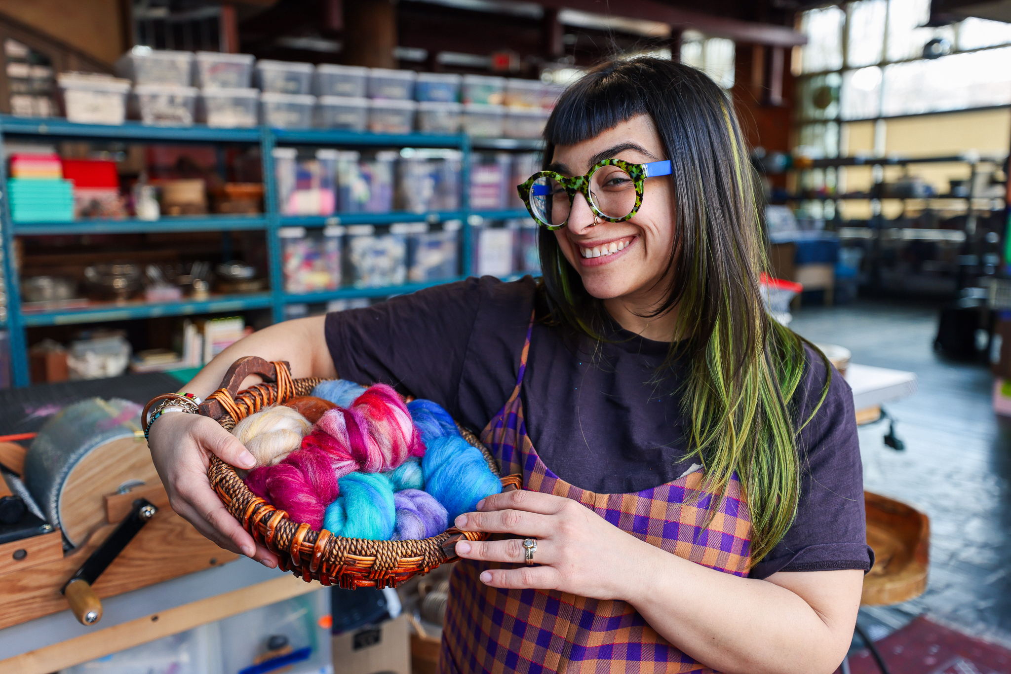 A smiling woman in brightly colored glasses and overalls stands in a large art studio, holding a basket full of colorful wool roving